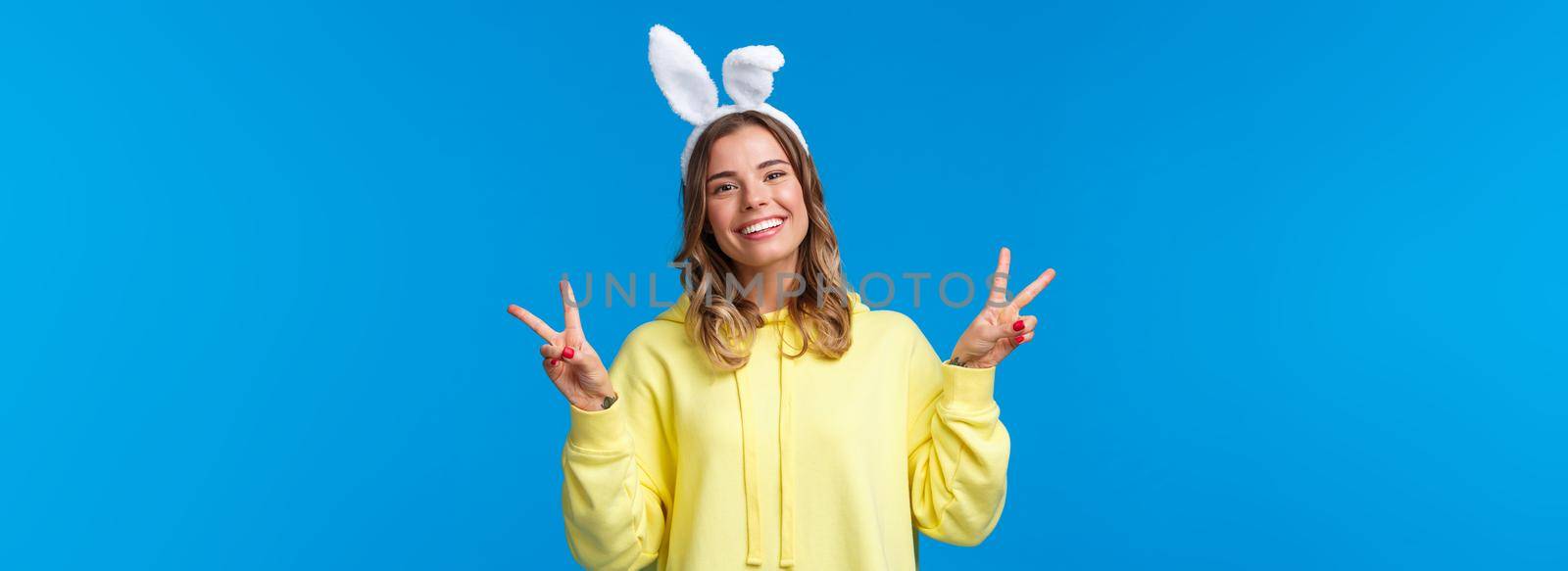 Holidays, traditions and celebration concept. Kawaii young blond girl in rabbit ears showing peace gesture and smiling, having fun, enjoying party, standing blue background.