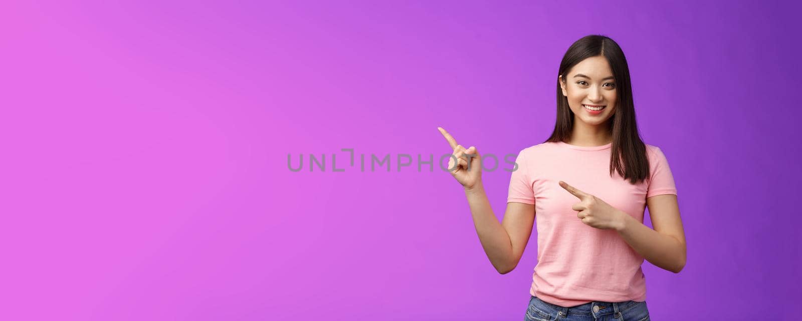 Friendly cute pleasant asian woman showing way, indicating interesting promo, pointing left smiling broadly, introduce advertisement product, stand purple background cheerful. Copy space