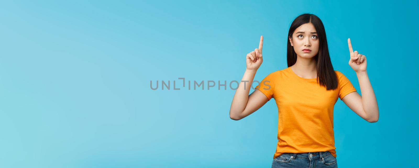 Upset gloomy cute asian young girl dark haircut frowning pointing looking up uneasy, feel regret cannot effort buy cool thing, stand blue background unsatisfied frustrated, wear yellow t-shirt.