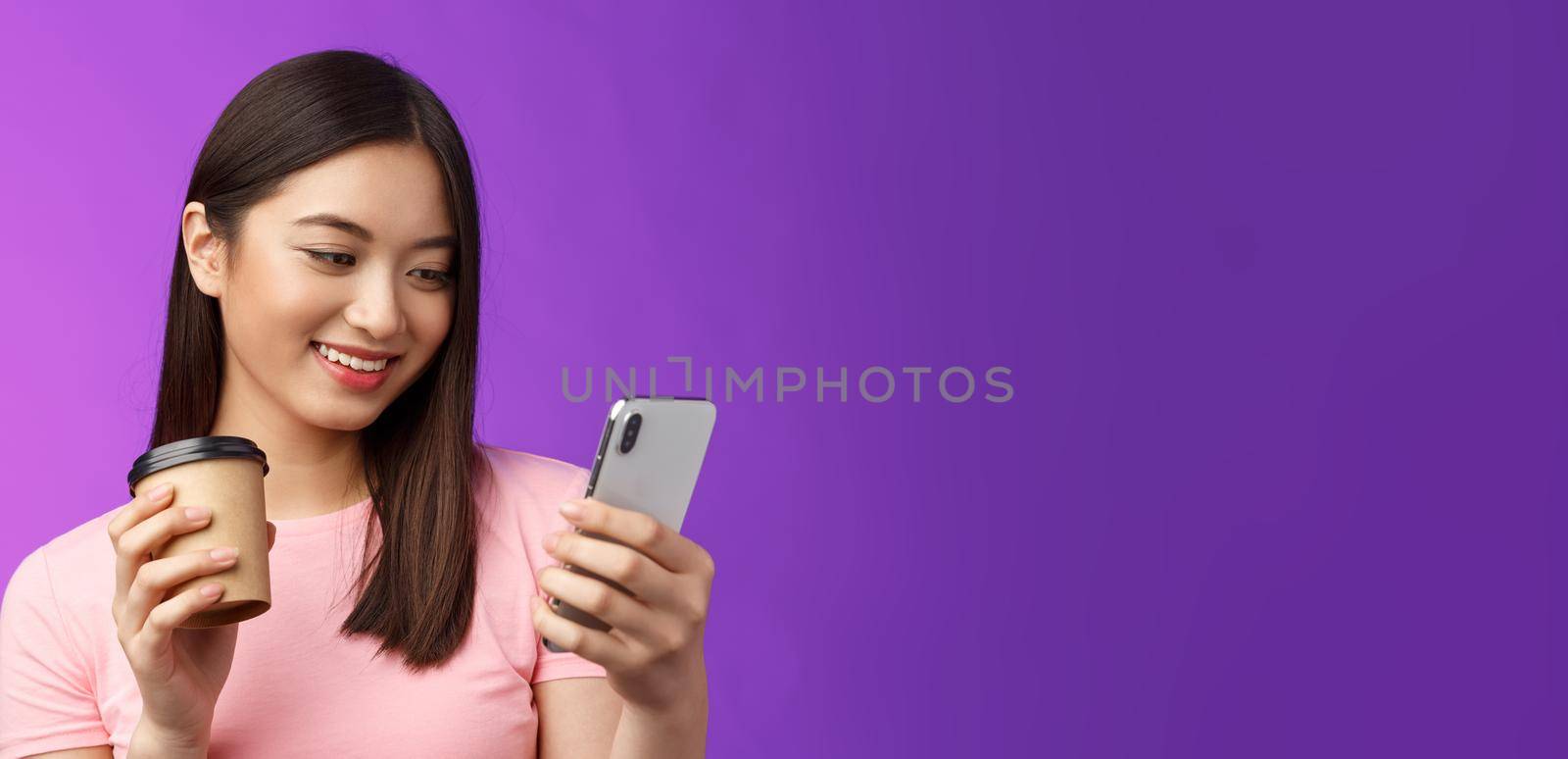 Close-up tender friendly asian female blogger checking social media drinking take-away coffee, smiling joyfully look telephone screen, reading interesting article, texting, purple background.