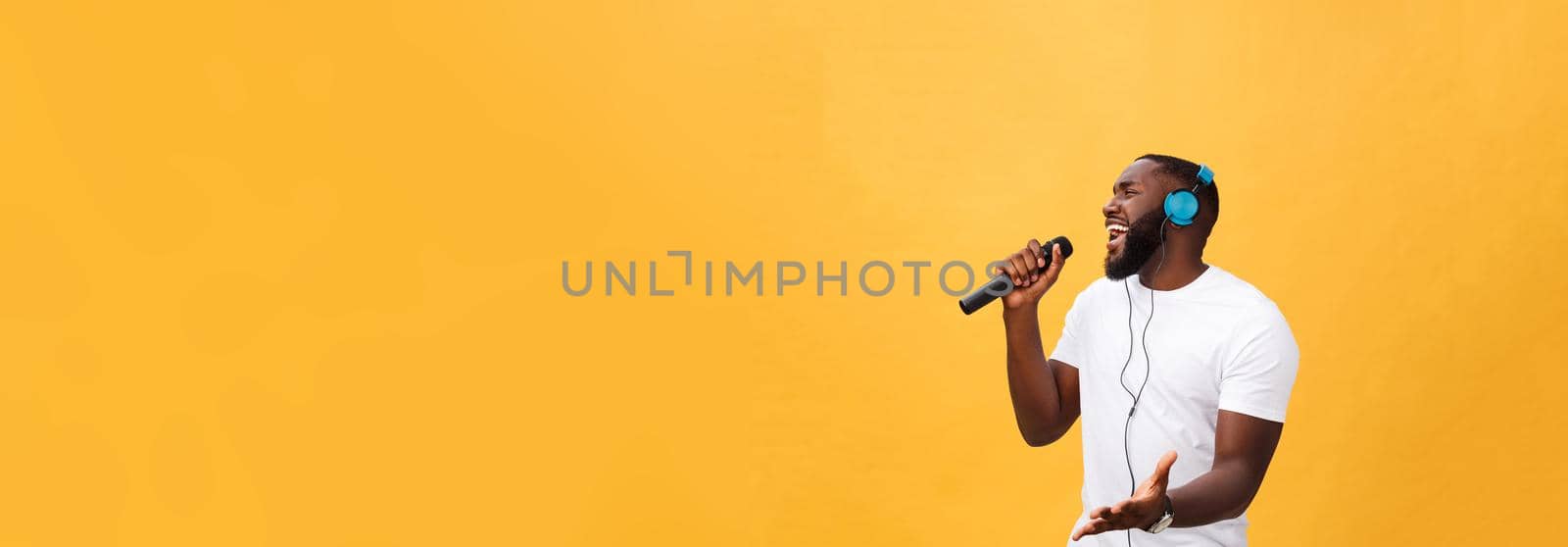 Portrait of cheerful positive chic. handsome african man holding microphone and having headphones on head listening music singing song enjoying weekend vacation isolated on yellow background.