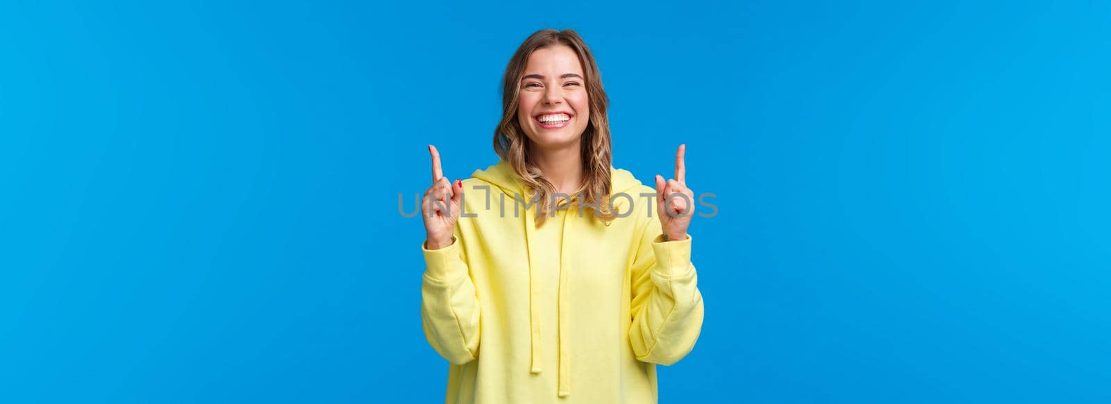 Cheerful happy smiling girl with blond hair in yellow hoodie, pointing fingers up and laughing carefree, recommend product, promo of subscribtion or company banner on blue background.