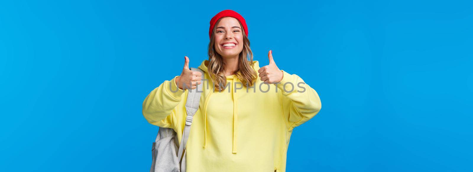 Sounds good, deal. Cheerful happy and smiling hipster girl in red beanie and yellow hoodie, approve idea, agree with friends picking where hang out after classes, hold backpack, blue background.