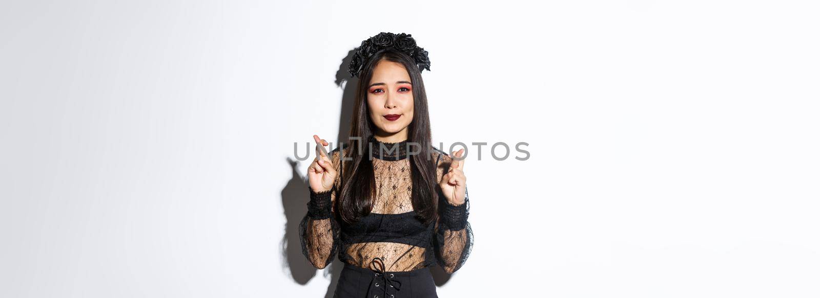 Image of hopeful asian girl in gothic lace dress and wreath making wish, cross fingers for good luck, looking wishful at camera, standing over white background.