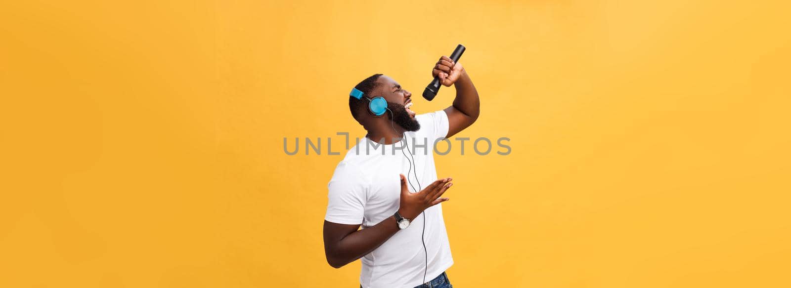 Portrait of cheerful positive chic. handsome african man holding microphone and having headphones on head listening music singing song enjoying weekend vacation isolated on yellow background.