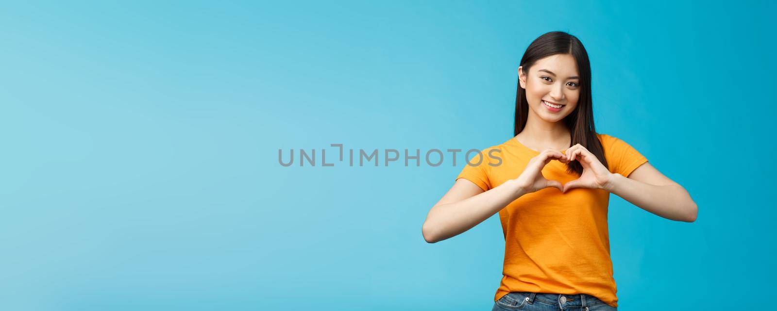 Lovely caring attractive asian girlfriend show heart sign cherish and value relationship, smiling broadly supportive, stand blue background wear yellow t-shirt, promote peace and happiness.