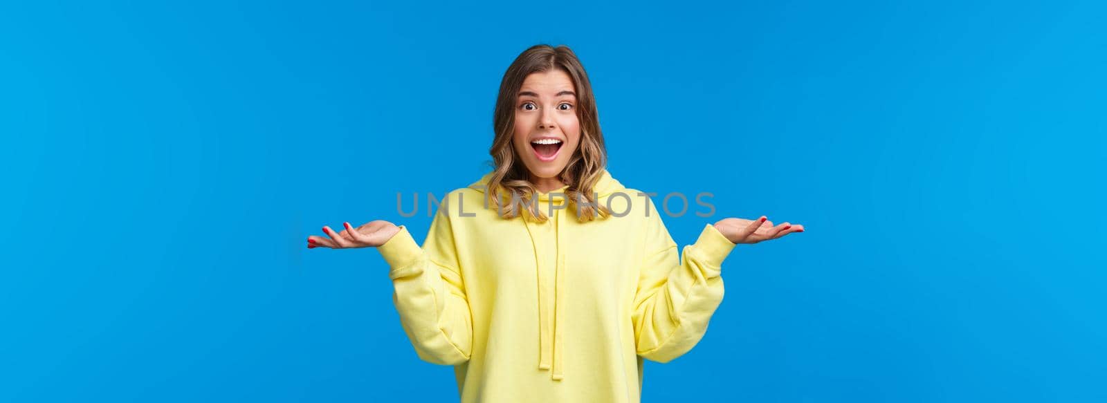 What nice surprise. Amused and happy cute blond caucasian woman spread hands sideways and shrugging smiling with excitement and disbelief, hear unexpected good news, blue background.