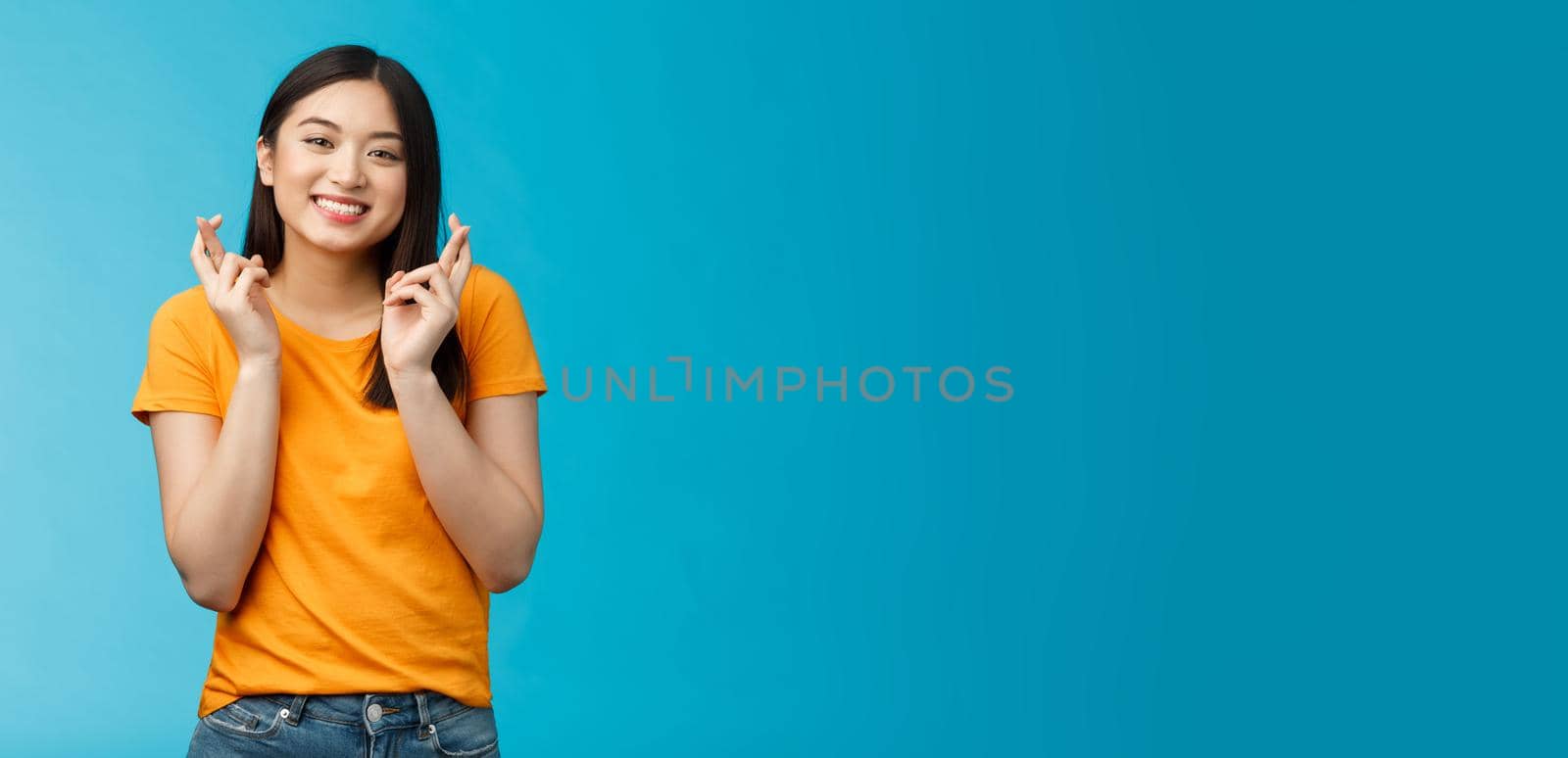 Cheerful smiling hopeful asian girl praying wish for good luck, cross fingers grinning toothy anticipate positive good news, stand blue background supplicating optimistic feelings hope win.