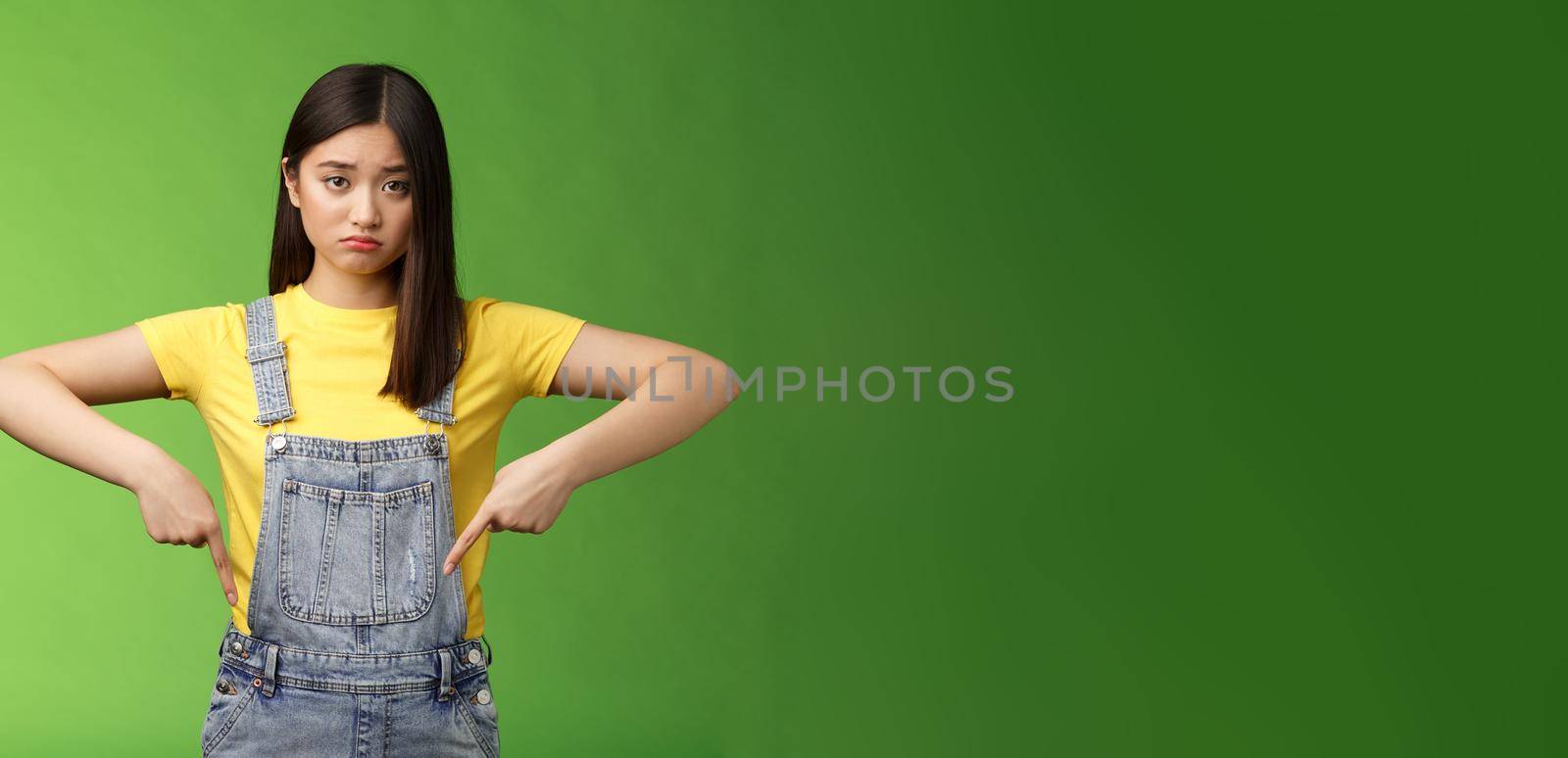 Sad asian girl in dispair feel upset, frowning sulking pessimistic, pointing down look camera pulling sorrow uneasy face, express sadness and frustration, stand green background. Copy space