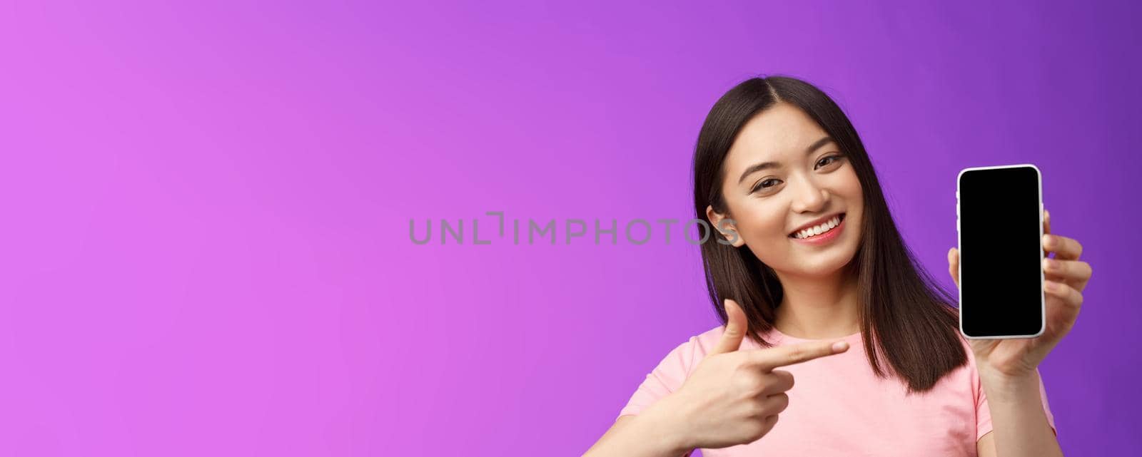 Close-up cheerful charismatic asian woman tilt head lovely, present app, show smartphone display introduce app, pointing telephone indicate index finger screen, stand purple background.