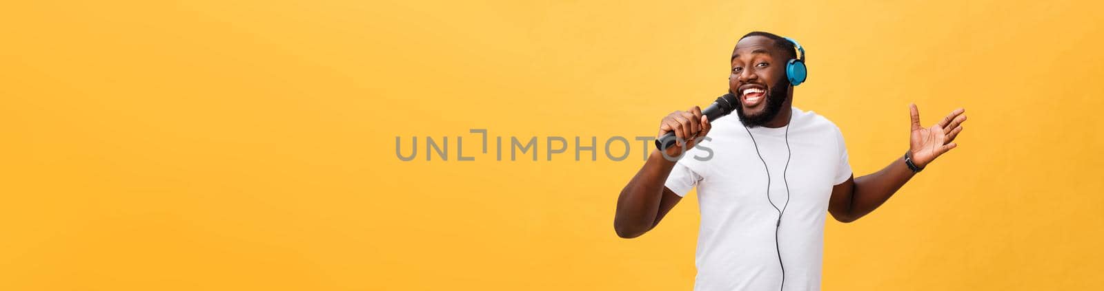 Portrait of cheerful positive chic. handsome african man holding microphone and having headphones on head listening music singing song enjoying weekend vacation isolated on yellow background.