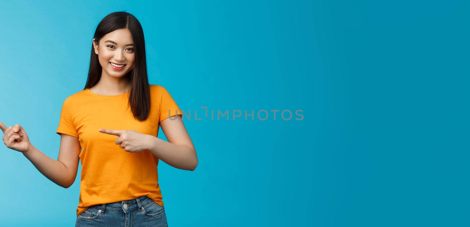 Friendly outgoing good-looking asian woman showing promo, pointing left indicating fingers advertisement, give advice, smiling confident, pleasantly help pick clothes, stand blue background.