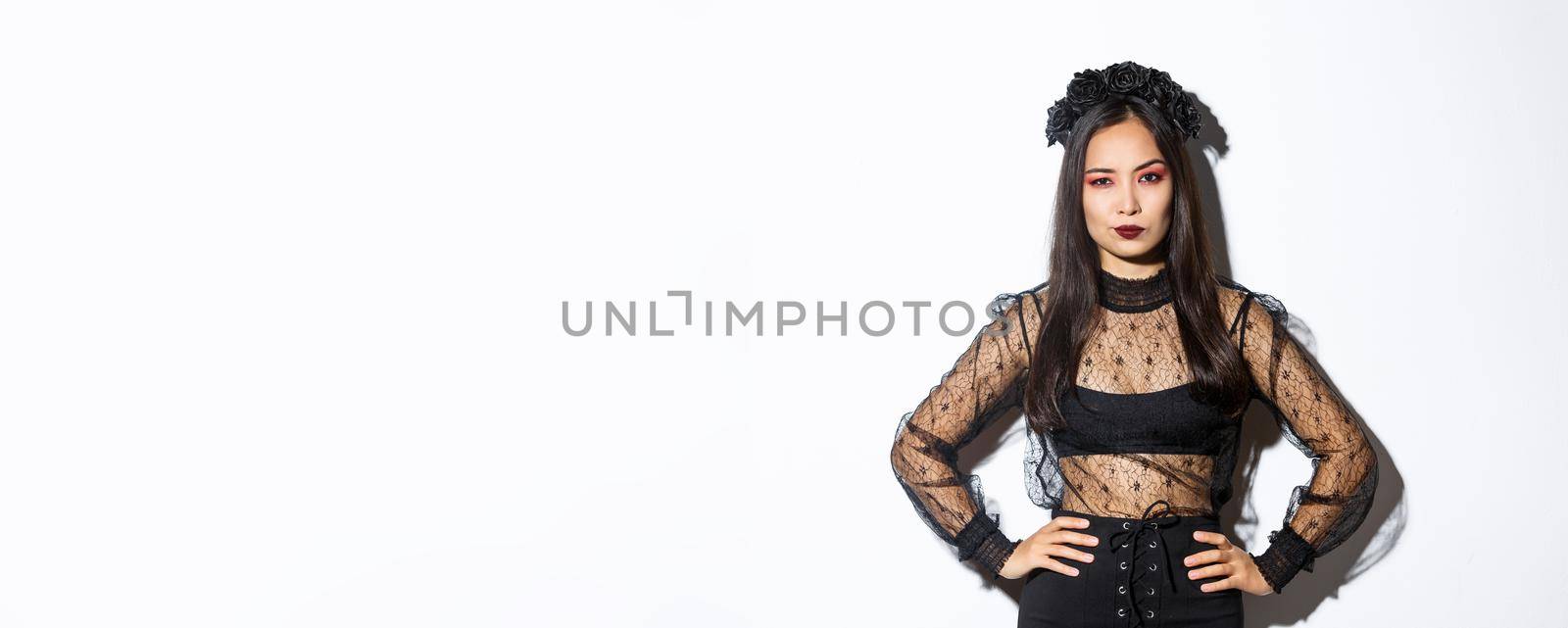 Attractive asian woman in halloween costume looking disappointed and skeptical. Female in black lace dress and wreath looking arrogant, trick or treat in witch outfit, standing white background.