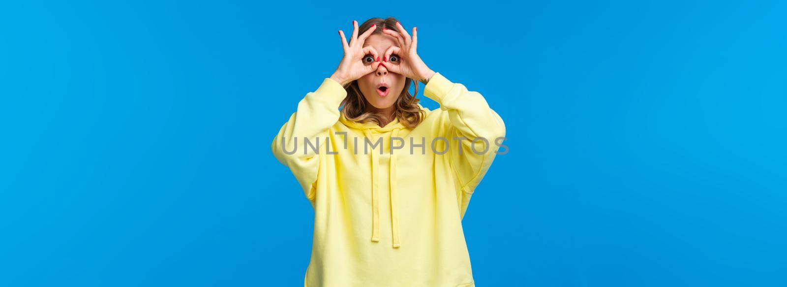 Wow interesting. Intrigued and surprised curious blond girl seeing something intriguing, open mouth astonished, look through hand-glasses and standing blue background.