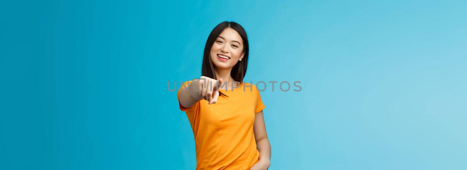 Lucky winner. Cheerful charismatic asian cute urban girl stand yellow t-shirt smiling friendly pointing finger camera choosing, picking person, inviting you team, stand happy blue background.