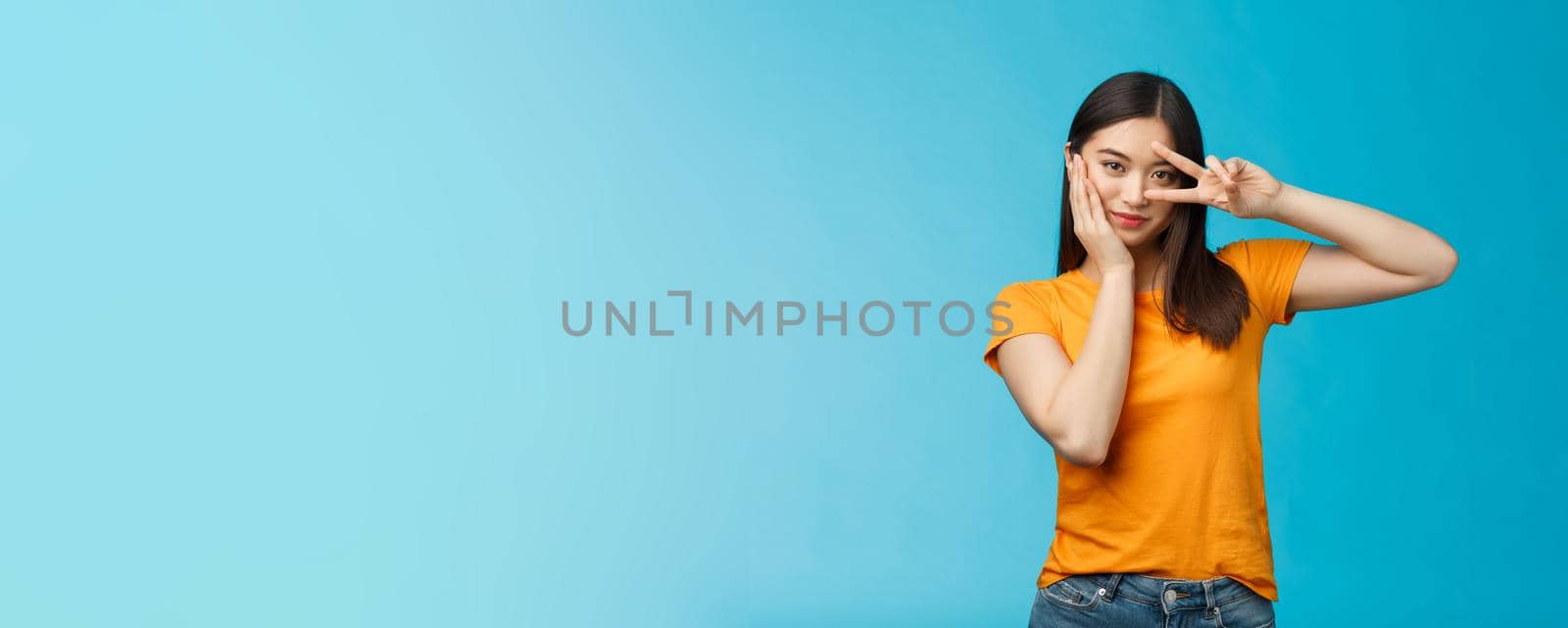 Sassy stylish good-looking asian assertive korean girl show peace victory sign on eye, touch cheek look empowered and motivated, standing cheeky coquettish blue background, wear yellow t-shirt.