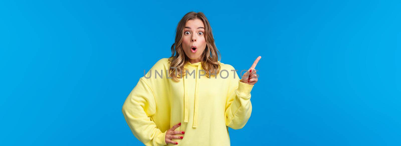 Wow incredible. Impressed and fascinated blond caucasian female pointing finger upper left corner and look camera astonished, telling about awesome event, standing blue background by Benzoix