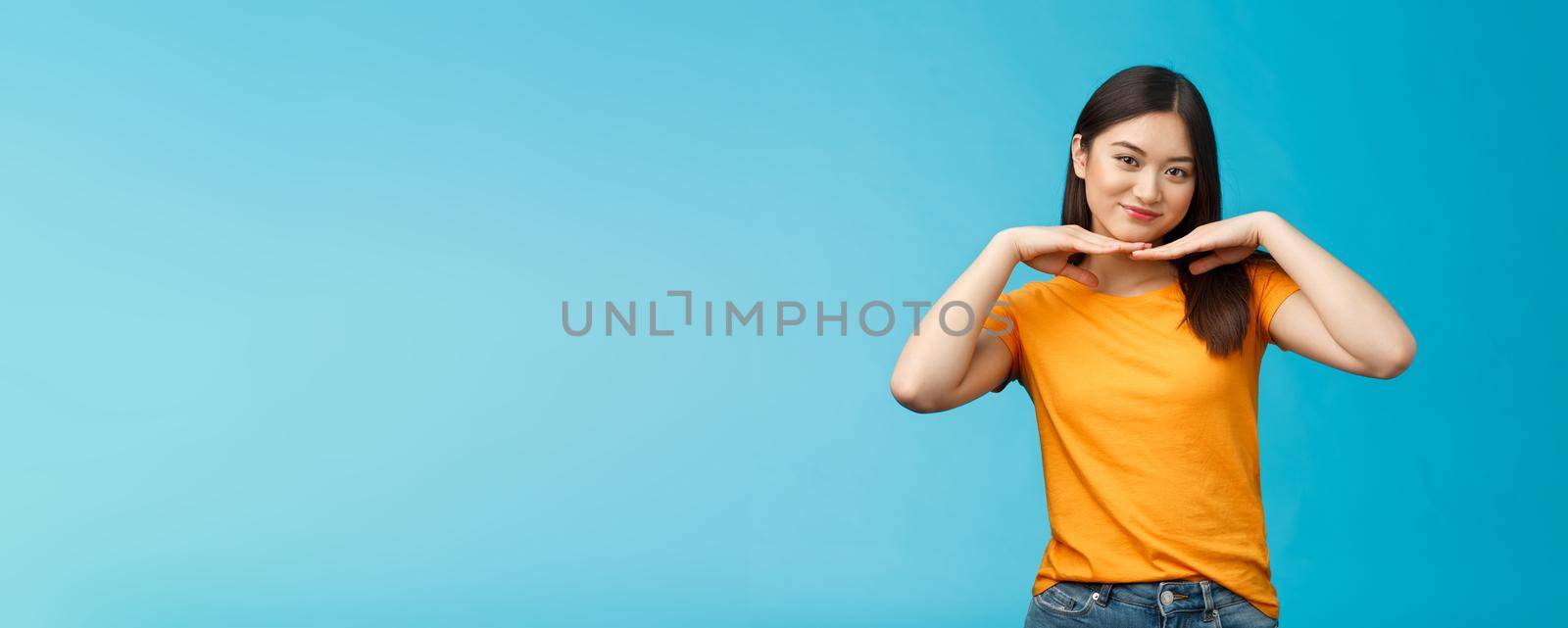 Sweet lovely tender asian girl brunette, tilt head smiling show clean pure face hold palms under jawline satisfied after applying skincare product stand blue background wear yellow t-shirt.