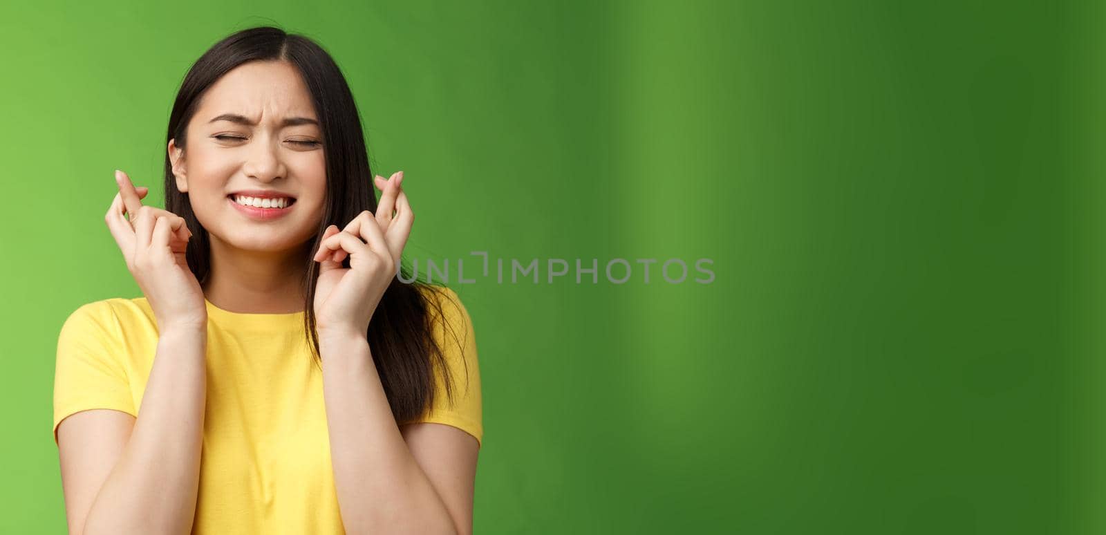 Nervous cute asian brunette girl eager win, worry important results, cross fingers good luck, praying fortune receive promotion, clench teeth close eyes supplicating, stand green background.