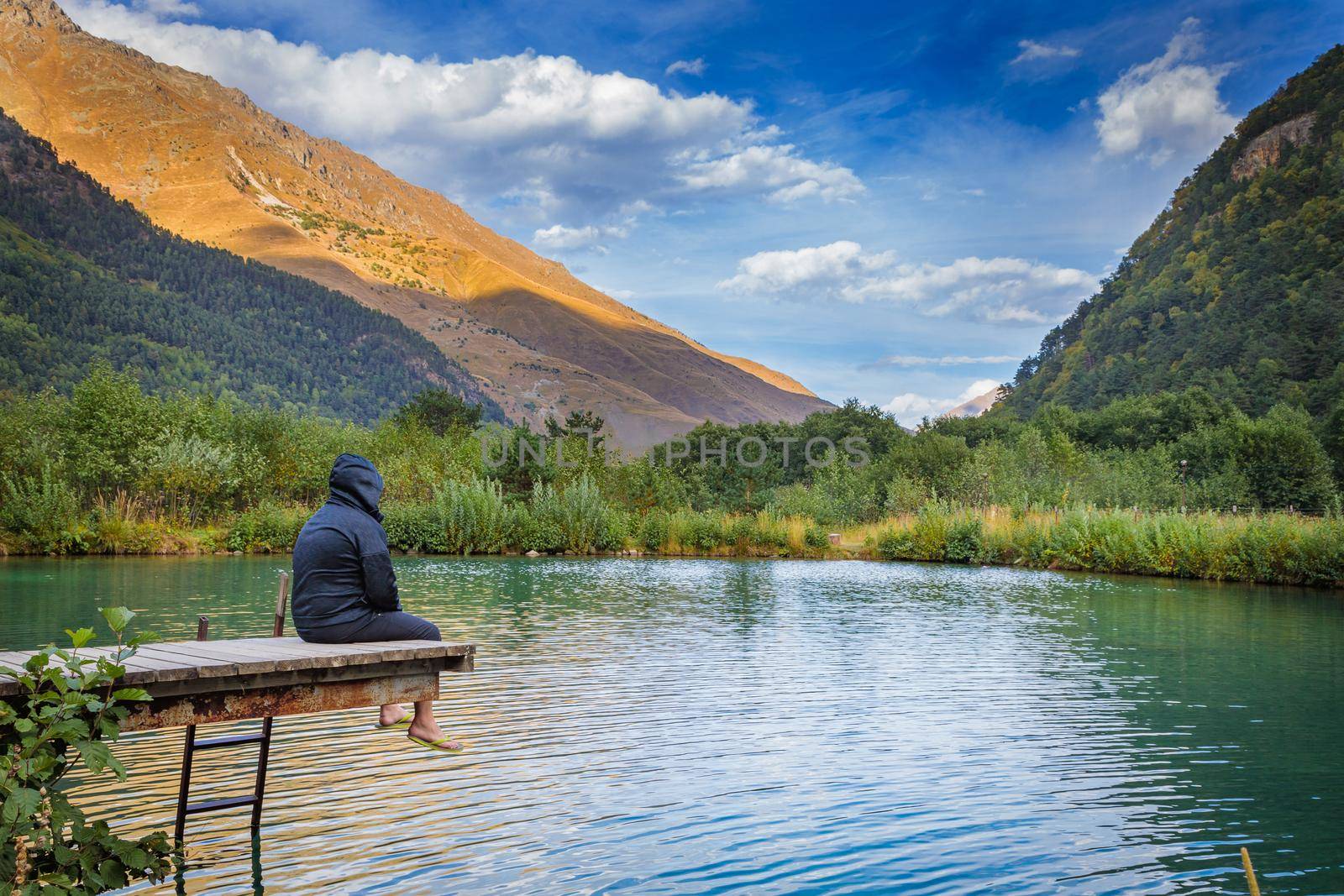 A man sits on a bridge near a mountain lake, enjoying the beauty of the mountains