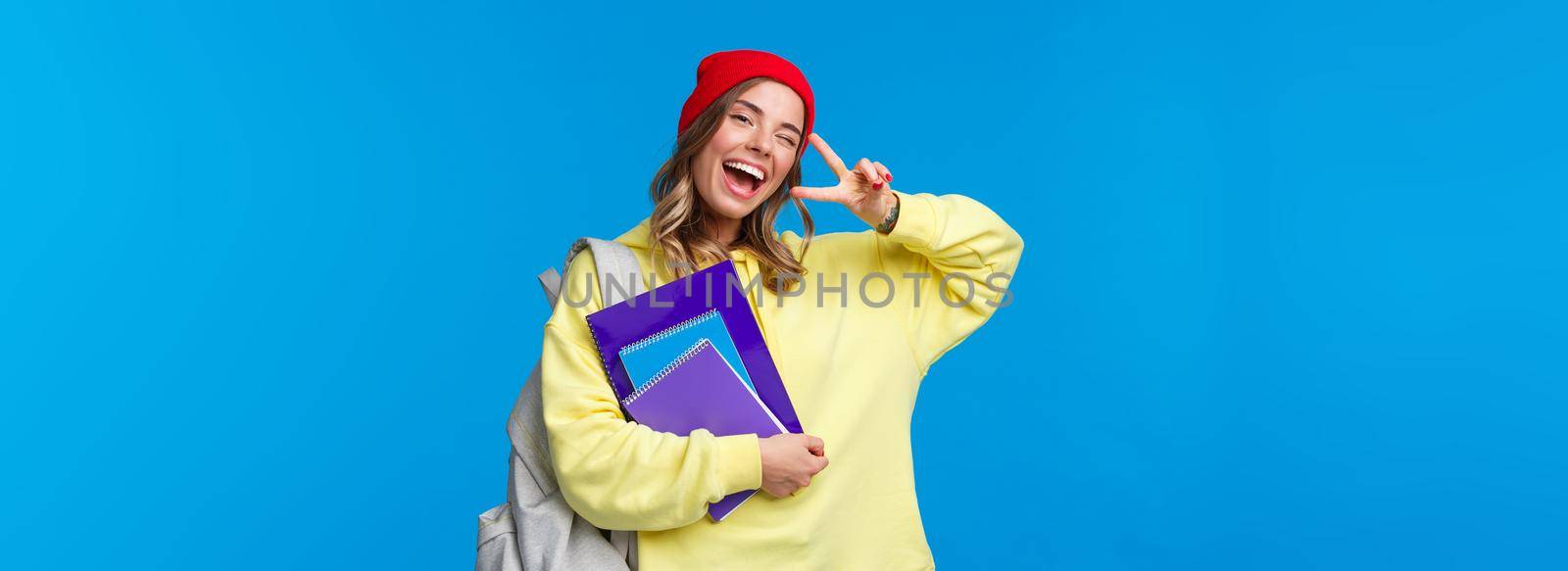 Education and learning concept. Cheerful smiling pretty female student showing peace gesture, enjoying college life, wink and gaze optimistic, studying hard, holding notebooks and backpack.