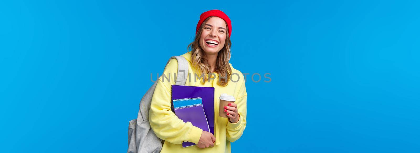 Cheerful happy female student having fun, enjoying university lifestyle, carry backpack and notebooks, drinking take-away coffee, talking to classmates, standing blue background joyful.
