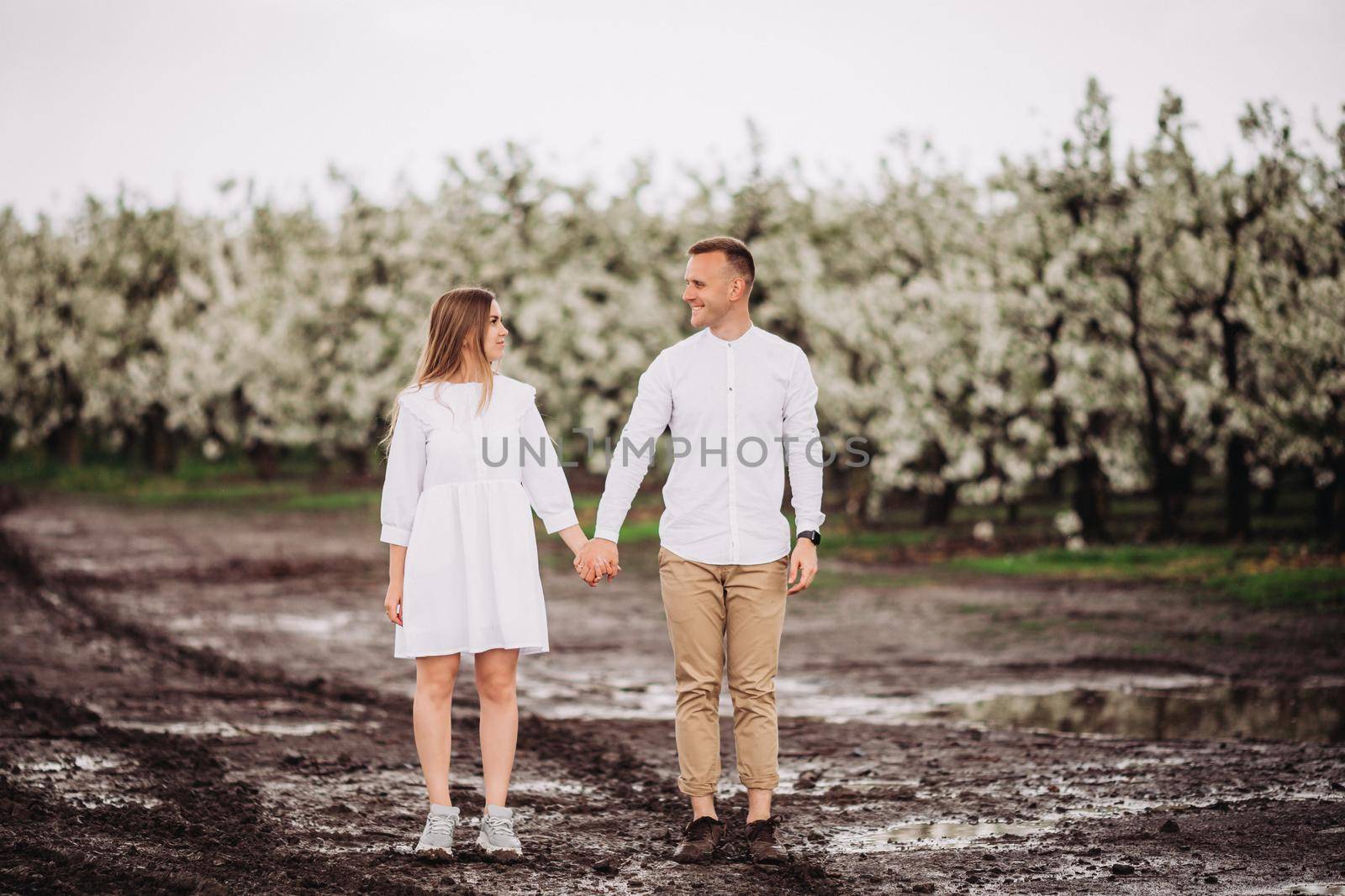 Happy family couple in the spring blooming apple orchard. Young couple in love enjoy each other while walking in the garden. Mud underfoot. The man holds the woman's hand. Family relationships by Dmitrytph