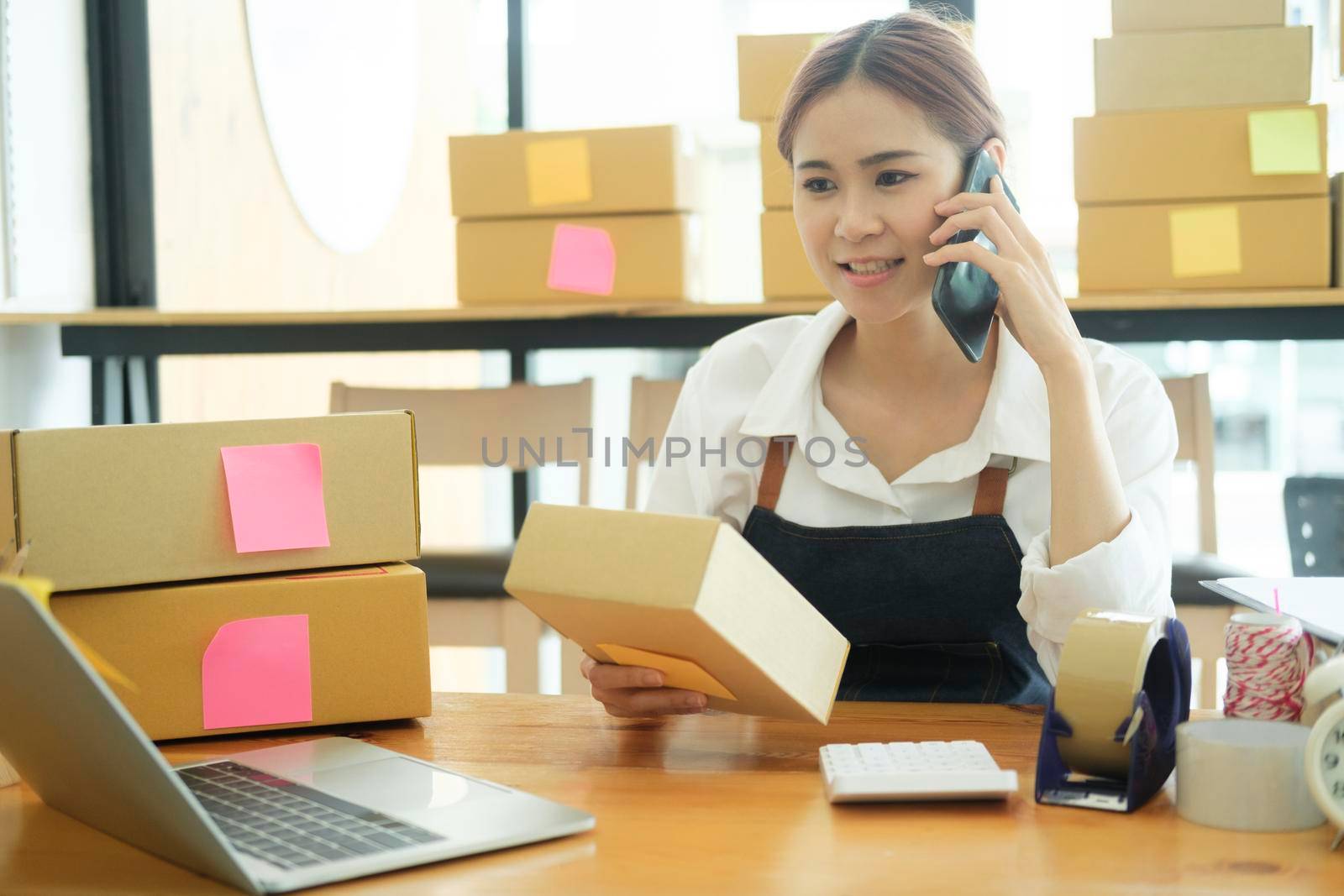 Female small online business owner talking on phone with clients to recieve and check order online, address preparing to pack order boxes for delivery. Online business concept.