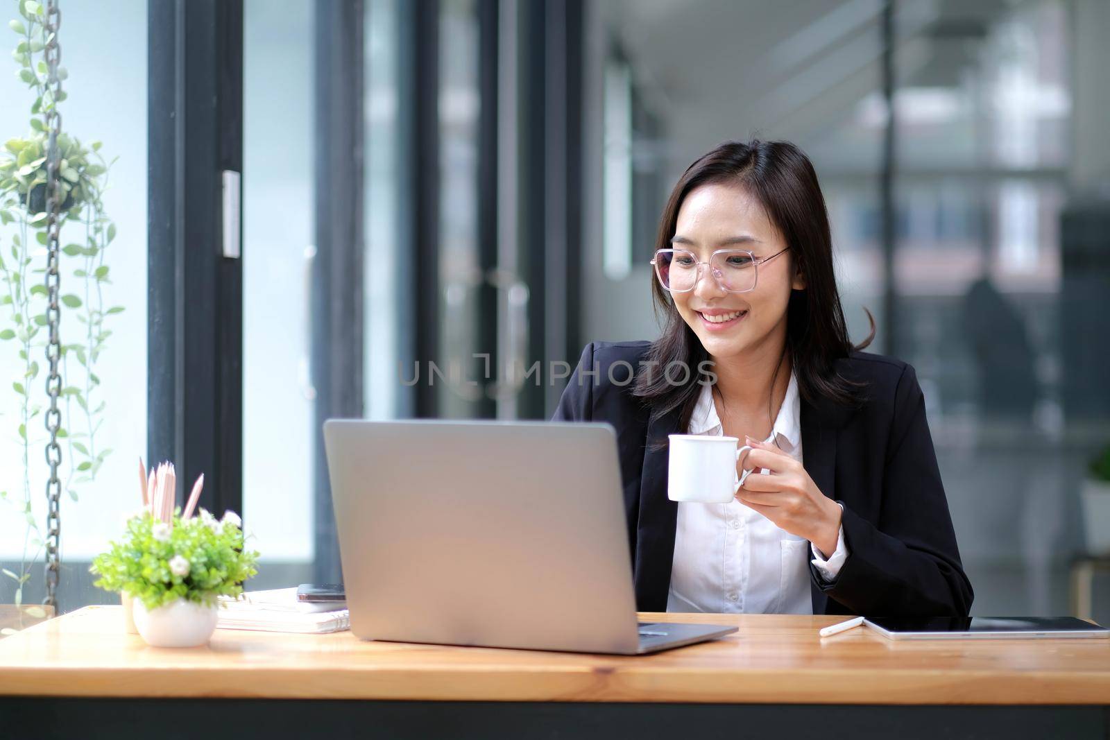 Professional asian businesswoman or female manager sits at her office desk, reading a business use laptop and drinking a morning coffee. by wichayada