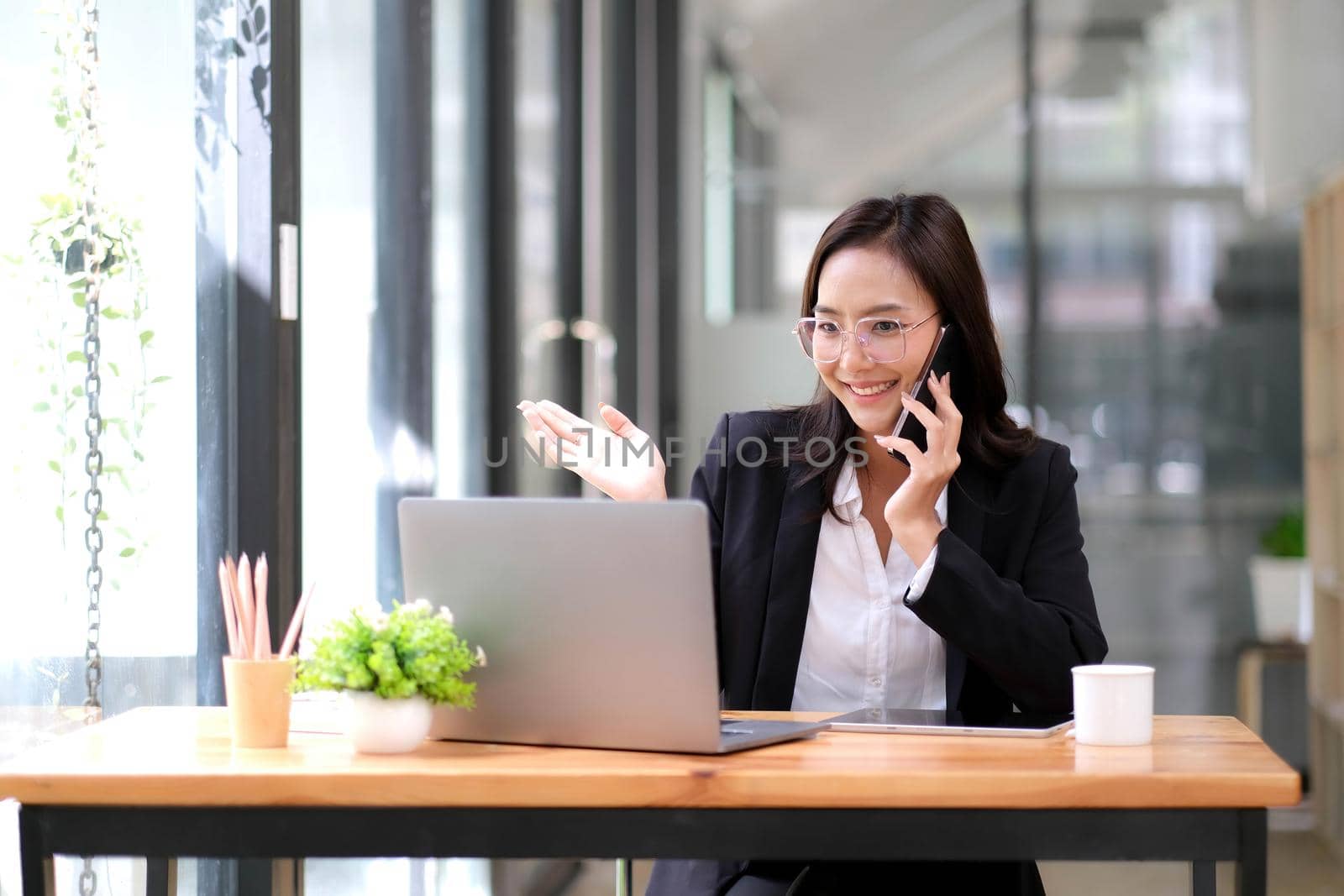 Happy businesswoman sitting at desk behind her laptop and talking with somebody on her mobile phone while working at office. by wichayada