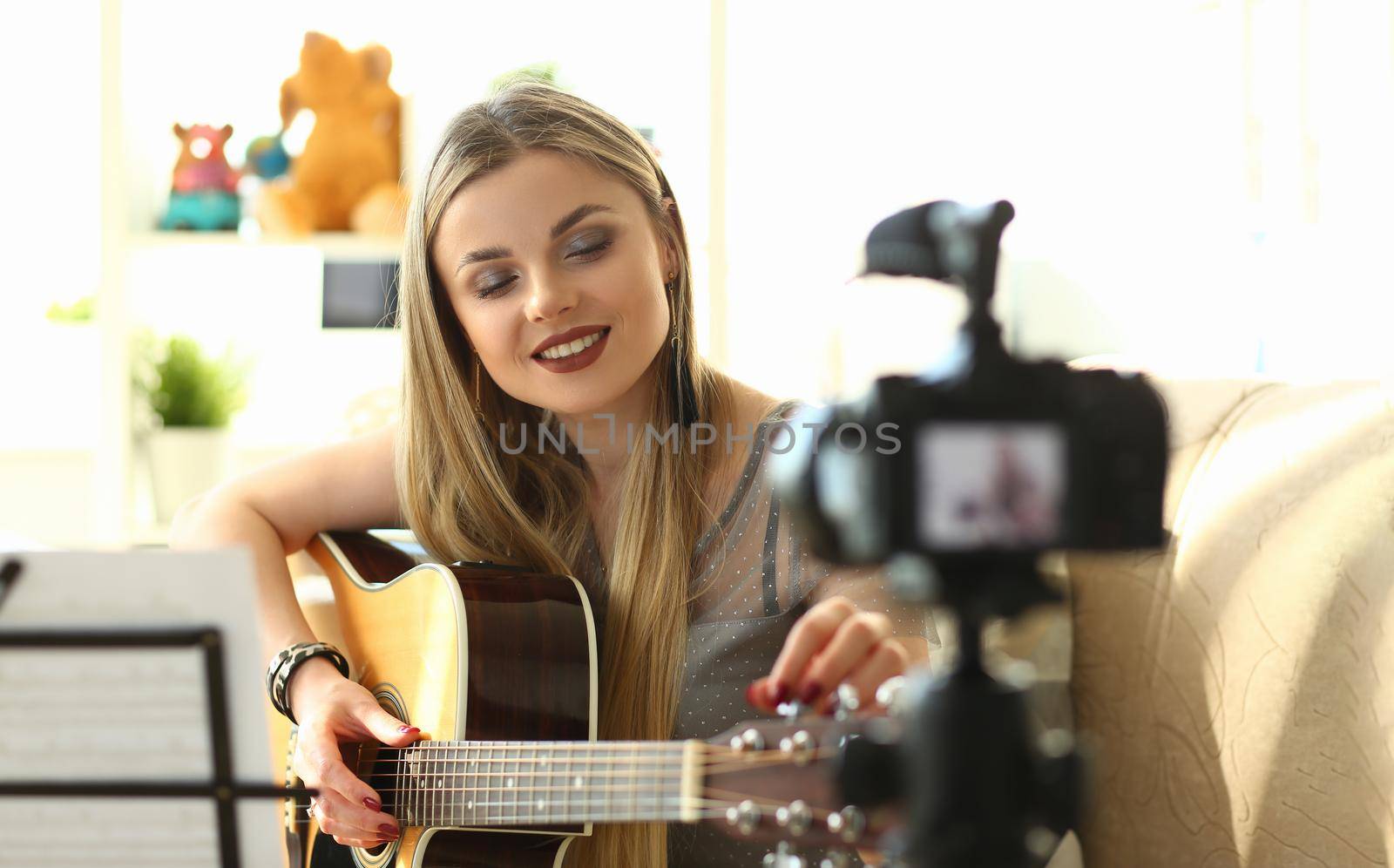 Young woman plays acoustic guitar and records video clips for social networks. Amateur musician creates content at home using modern camera concept