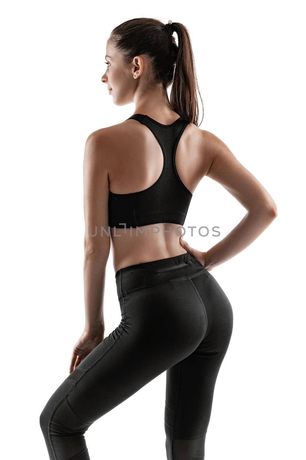 Strong brunette maiden with a ponytail, in black leggings, top and sneakers is posing standing back to the camera isolated on white background. Chic muscular body, fitness, gym, healthy lifestyle concept. Close-up portrait.