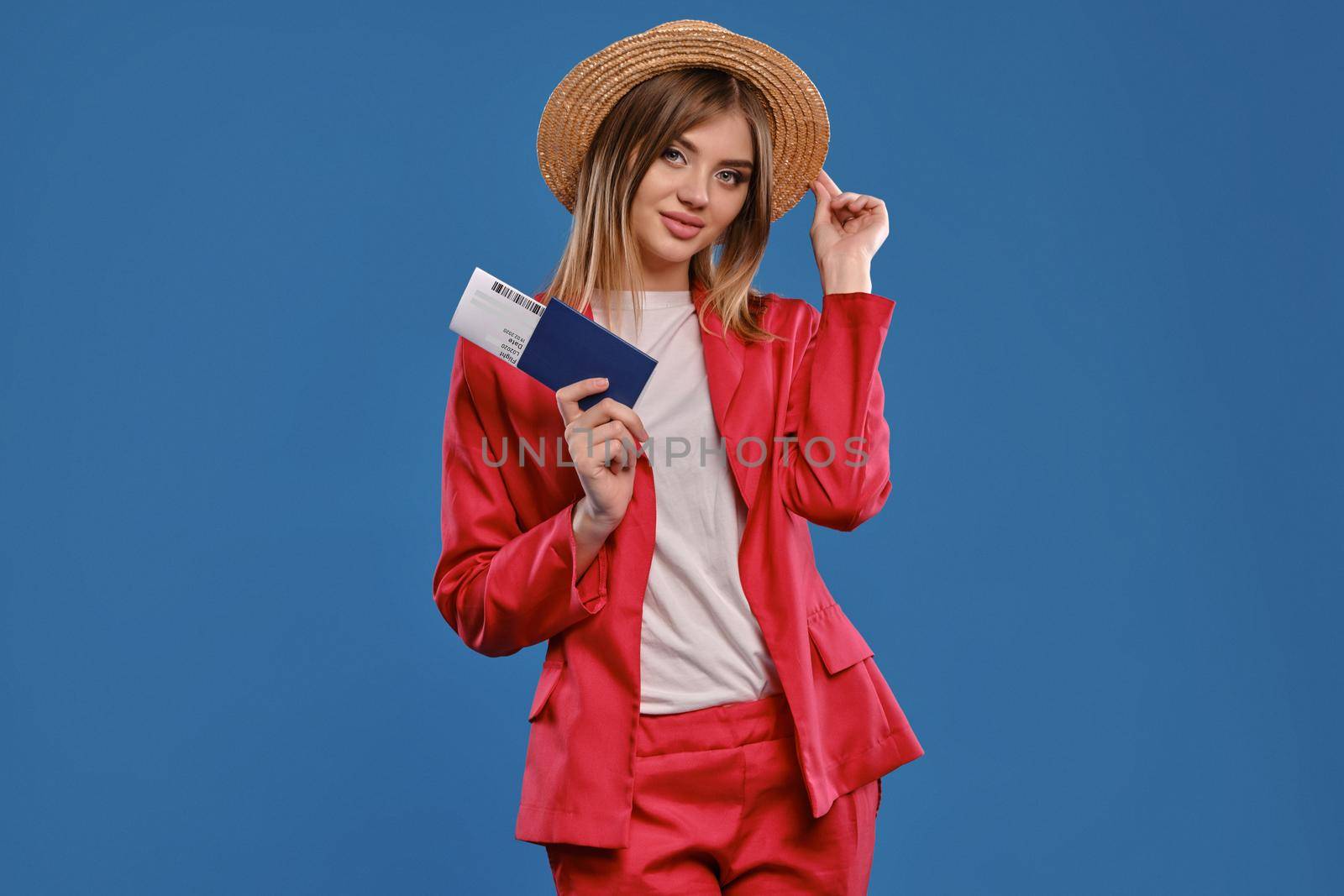 Alluring blonde model in straw hat, white blouse and red pantsuit. She is smiling, touching headdress, holding passport and ticket, posing on blue background. Travelling concept. Close-up, copy space