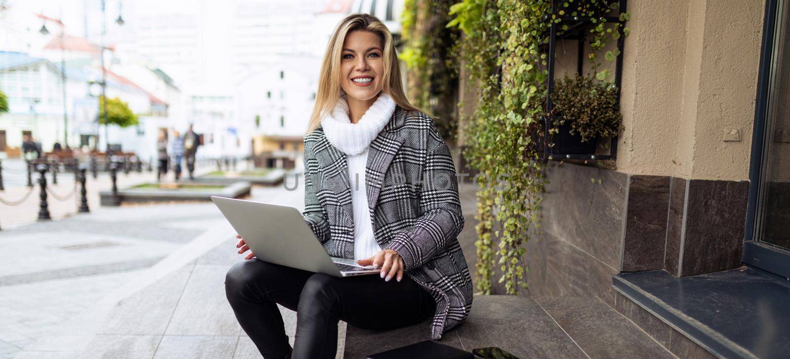 adult woman working on laptop outside office sitting on steps smiling looking at camera.