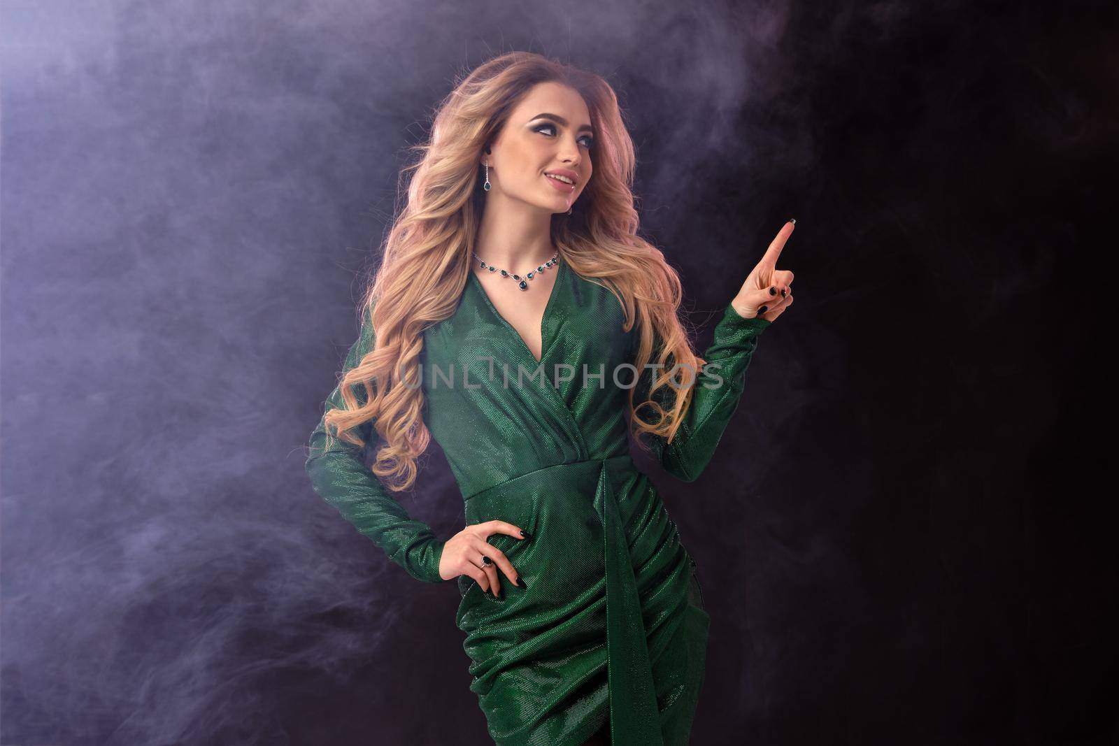 Alluring blonde female in green stylish dress and jewelry. Smiling, pointing at something, posing on black smoky background. Template, mockup for your advertising and design. Close up, copy space