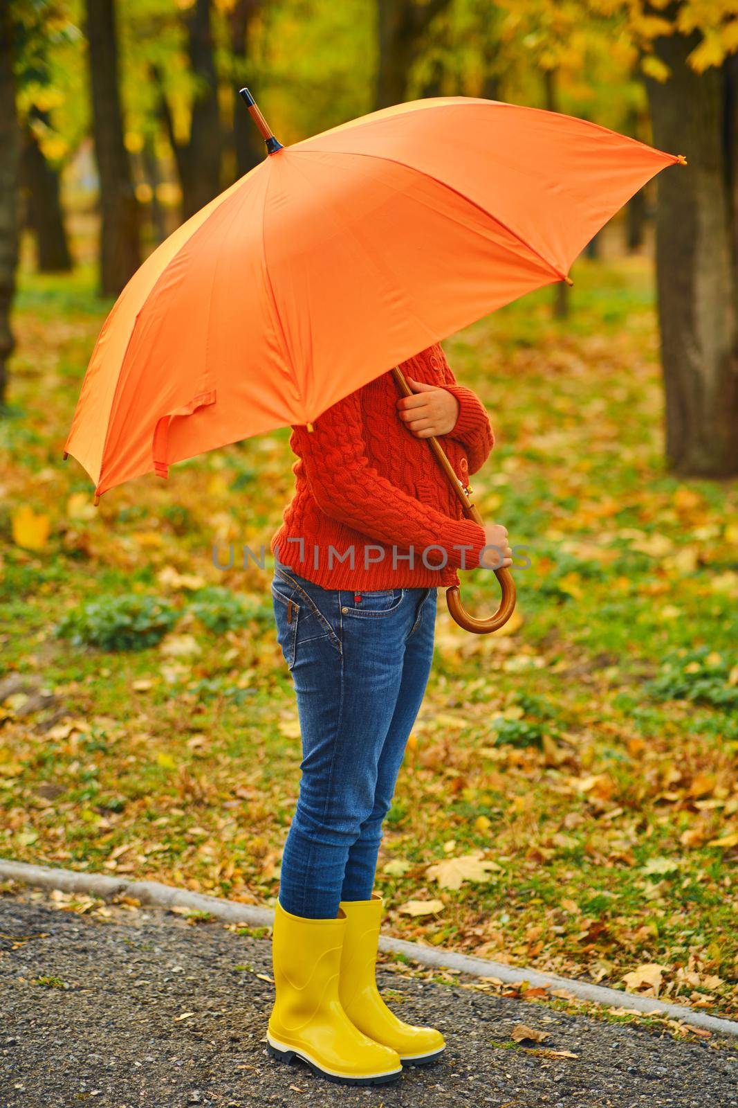 Funny unrecognizable kid in rubber boots under an orange umbrella outdoors in Autumn park