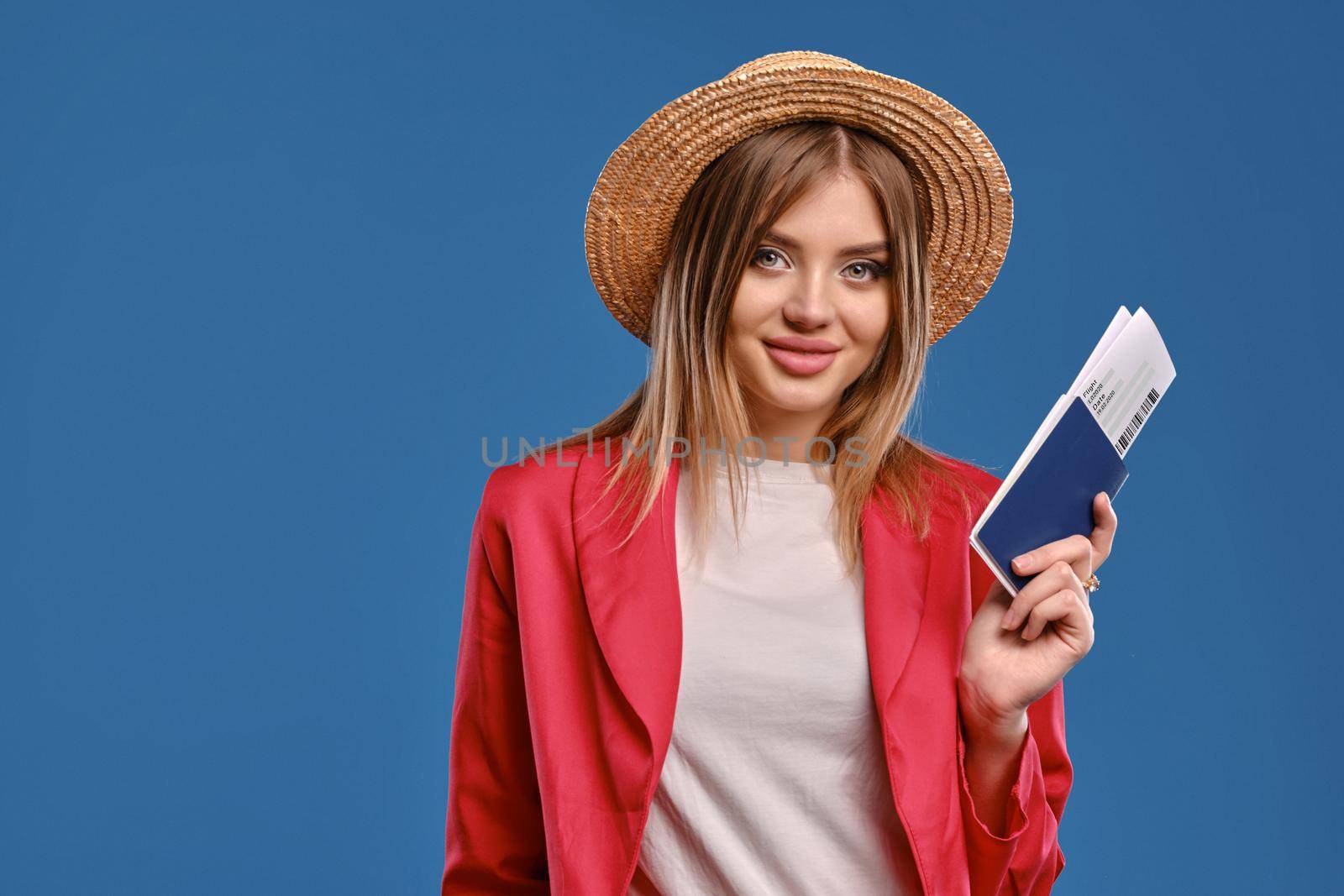 Pretty, blonde girl in straw hat, white blouse and red pantsuit. She is smiling, showing her passport and ticket, posing against blue studio background. Travelling concept. Close-up, copy space