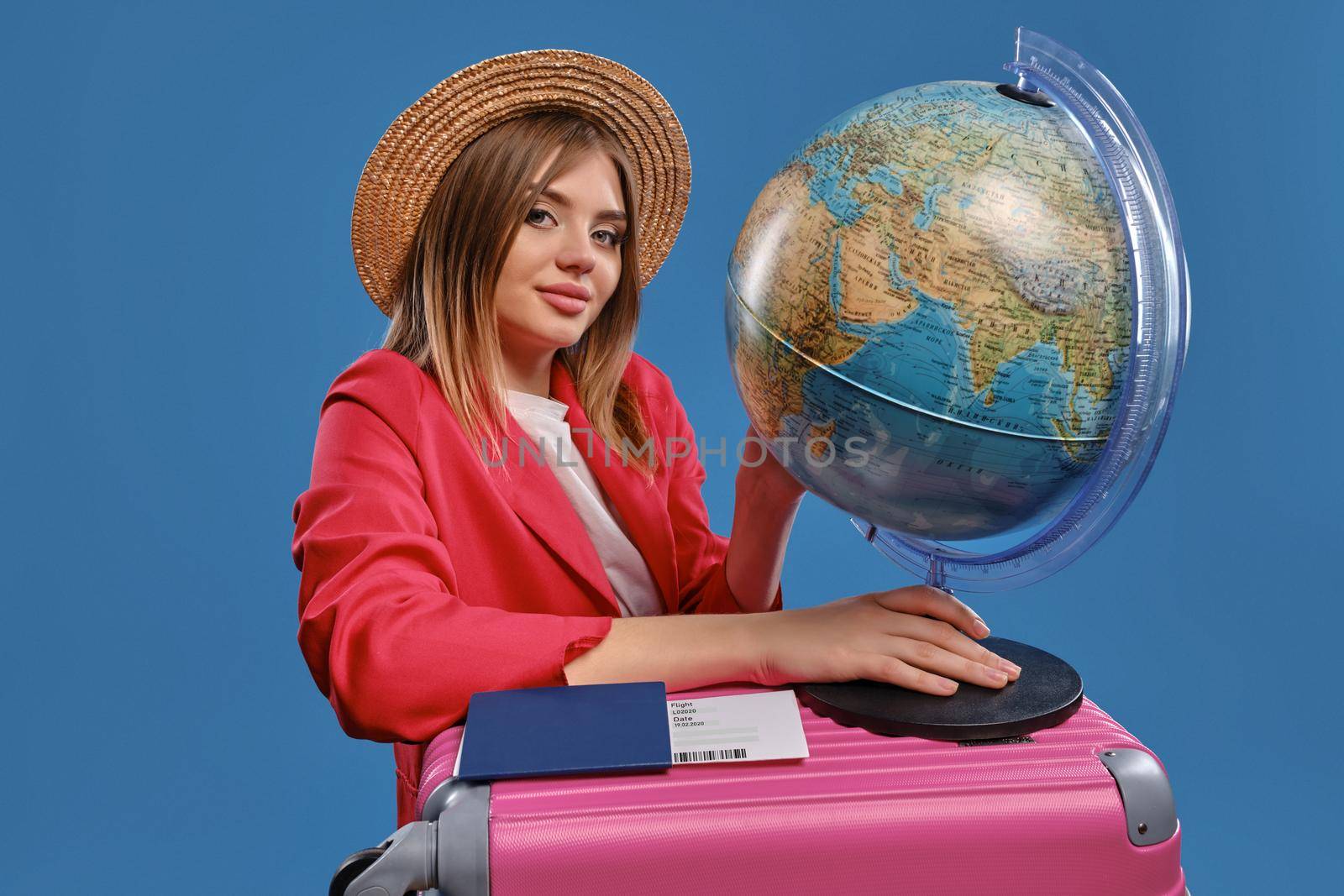 Elegant blonde girl in straw hat, white blouse, red jacket. She smiling, holding globe standing on pink suitcase, passport and ticket are nearby, posing on blue background. Close-up, copy space
