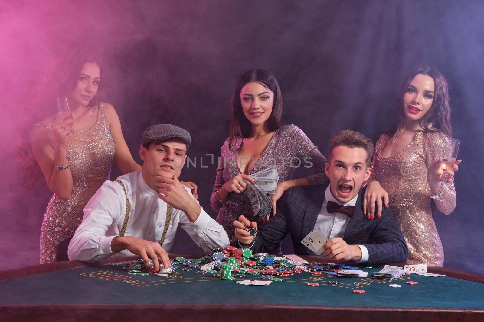 Buddies playing poker at casino, at table with stacks of chips, money, cards on it. Happy about win, drinking champagne, smiling. Black, smoke background, colorful backlights. Gambling. Close-up.