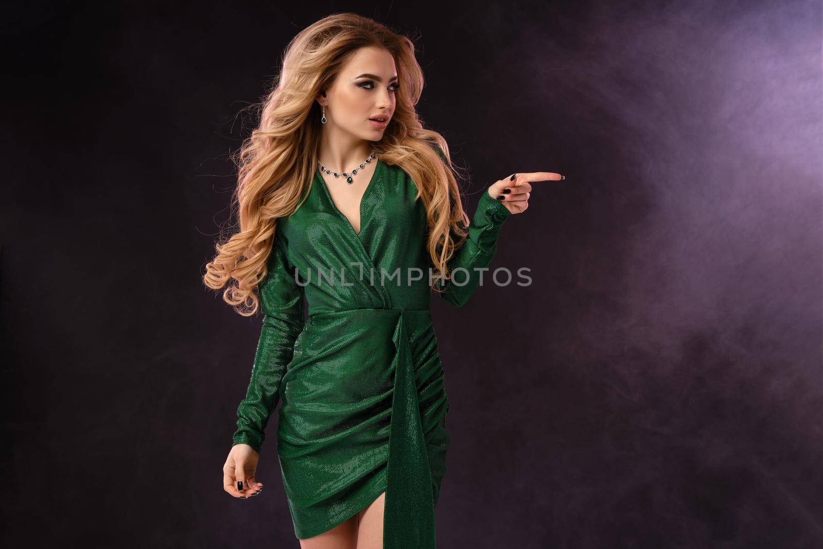 Blonde lady, make-up, in green stylish dress and jewelry. Pointing at something by forefinger, posing on black smoky background. Template, mockup for your advertising and design. Close up, copy space