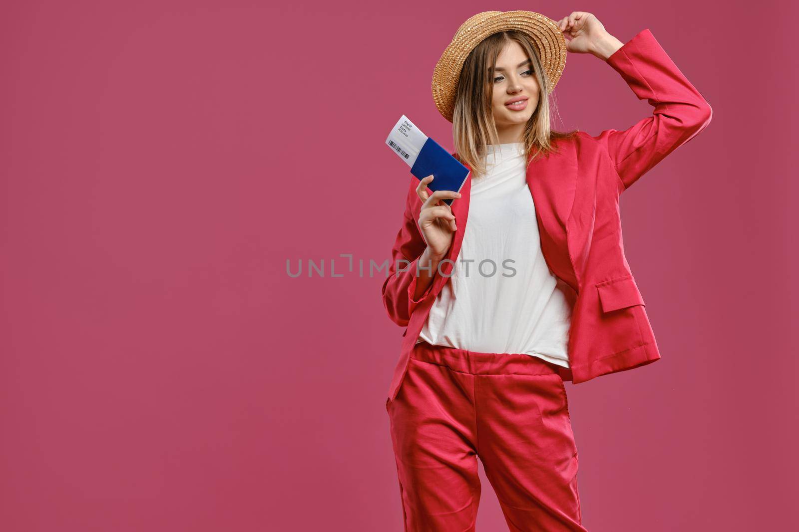 Charming blonde female in straw hat, white blouse and red pantsuit. Smiling, touching headdress, holding passport and ticket while posing on pink background. Travelling concept. Close-up, copy space