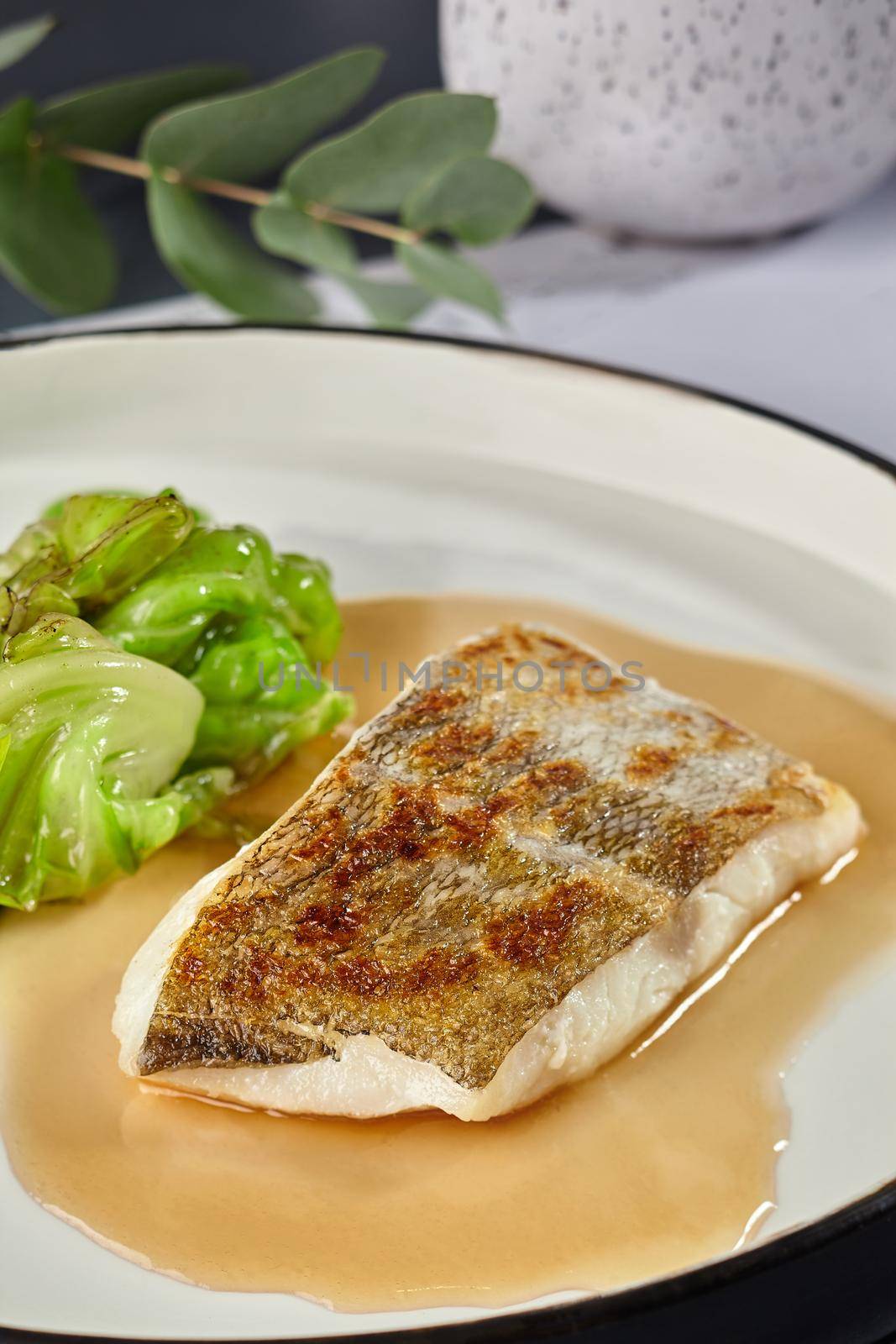 Yummy fish cooked in an oven and garnished with sauce and cabbage, served on a white plate with black edging, dark table, cutlery. Selective focus. Top view, restaurant cooking concept.