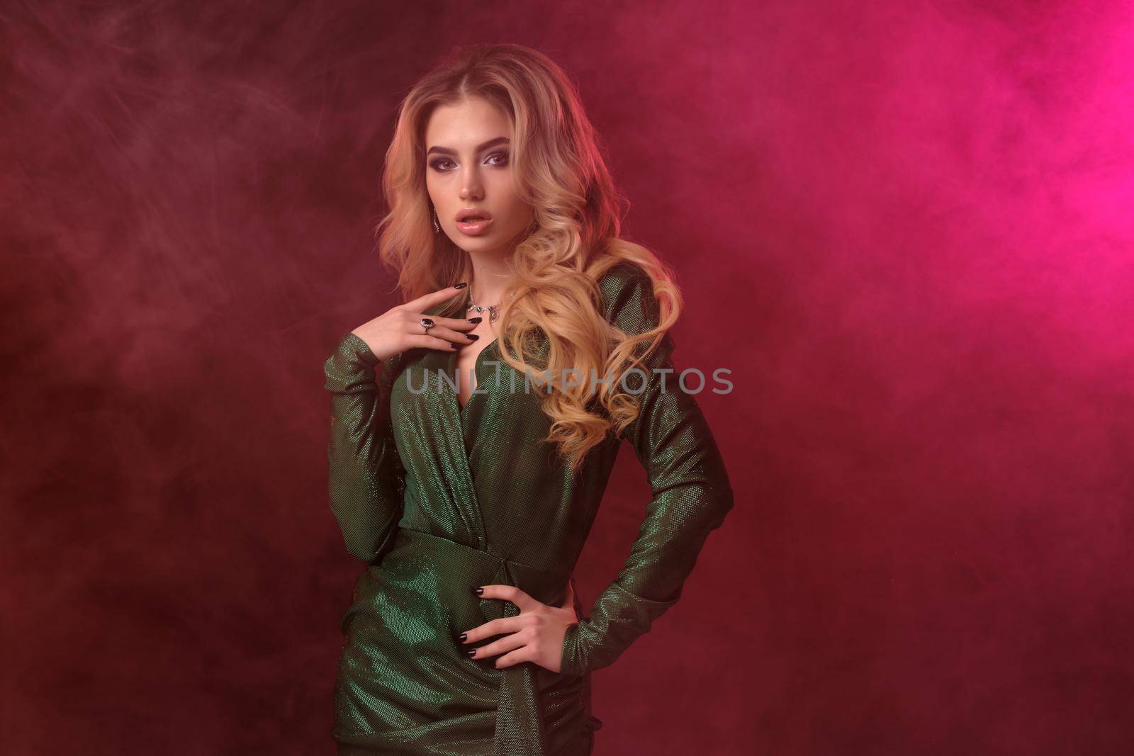 Alluring, blonde curly female in green stylish dress and jewelry. She has put her hand on waist, looking at you, posing against colorful smoky studio background. Fashion and beauty concept. Close up