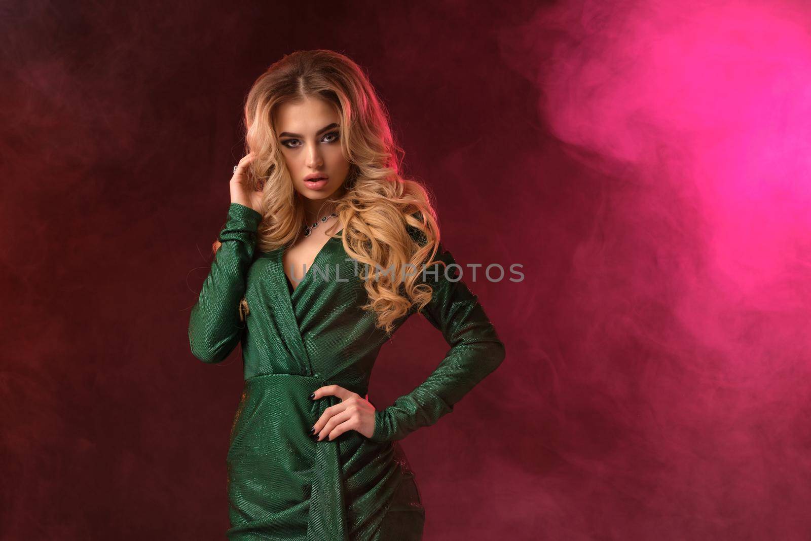 Attractive blonde woman with bright make-up, in green stylish dress and jewelry. She is touching her hair, hand on waist, posing against colorful smoky studio background. Fashion and beauty. Close-up.