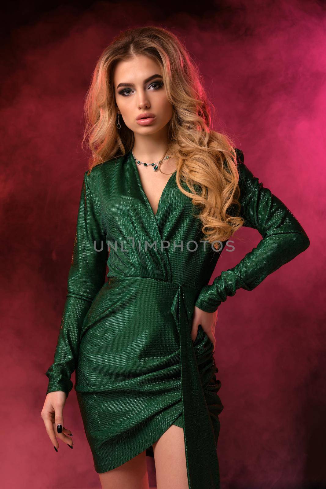 Gorgeous blonde curly woman with bright make-up, in green stylish dress and jewelry. She has put her hand on waist, posing against colorful smoky studio background. Fashion and beauty. Close up