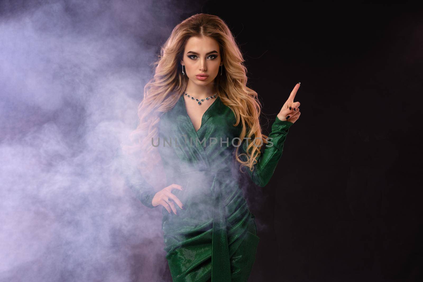 Attractive blonde girl, make-up, in green stylish dress and jewelry. Pointing at something, posing on black smoky background. Template, mockup for your advertising and design. Close up, copy space