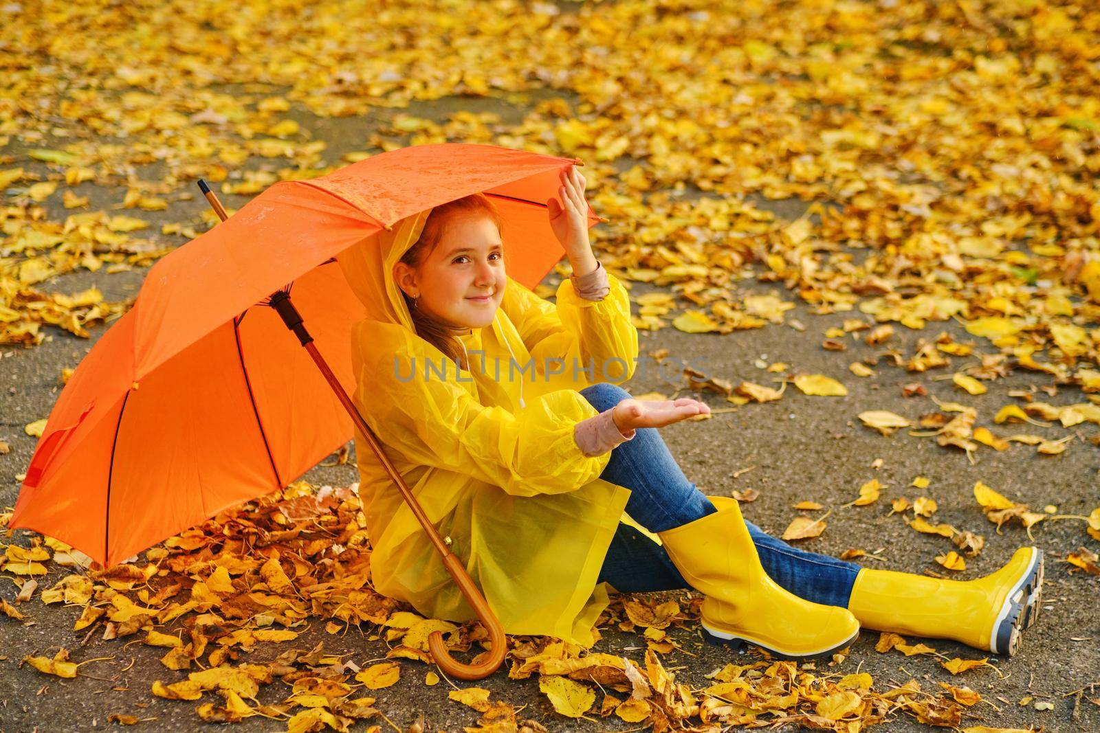 Happy kid Sits under an orange umbrella in the autumn park catching rain drops