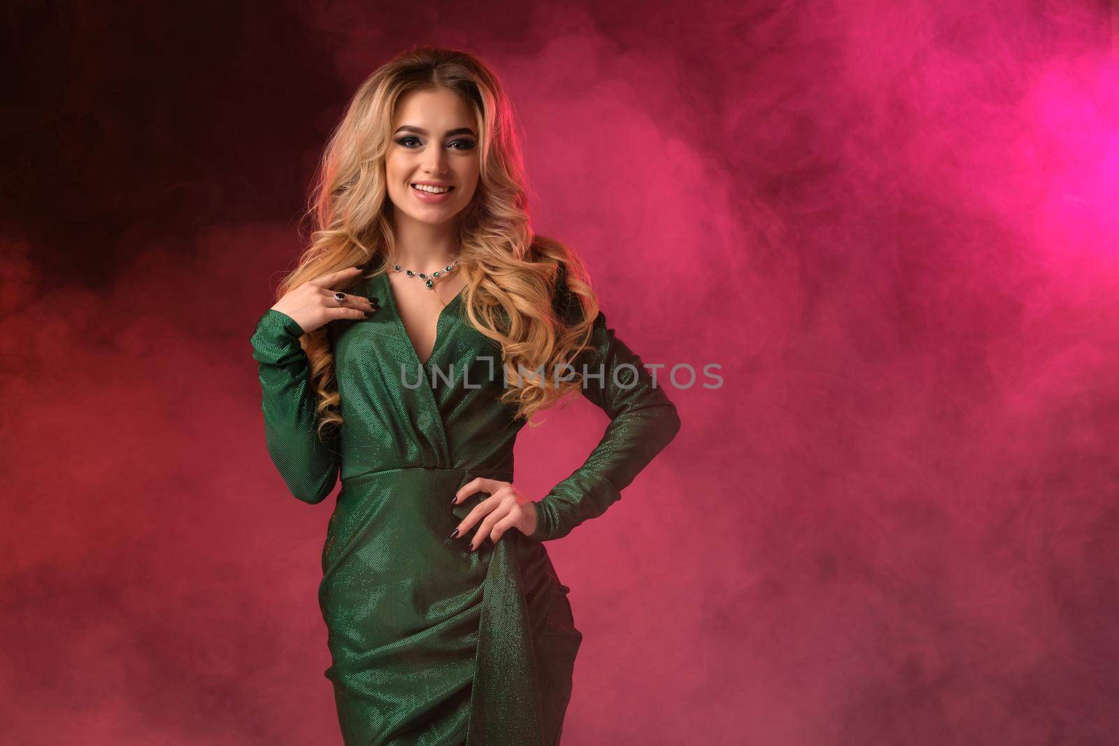 Attractive, blonde curly female in green stylish dress and jewelry. She put her hand on waist, smiling, looking at you, posing against colorful smoky studio background. Fashion and beauty. Close up