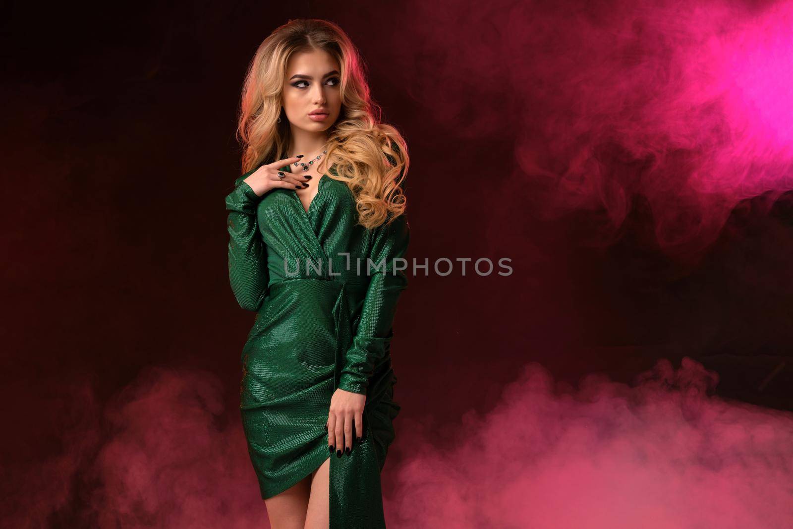 Gorgeous blonde curly girl with bright make-up, in green stylish dress and jewelry. She has put her hand on chest, posing against colorful smoky studio background. Fashion and beauty. Close up