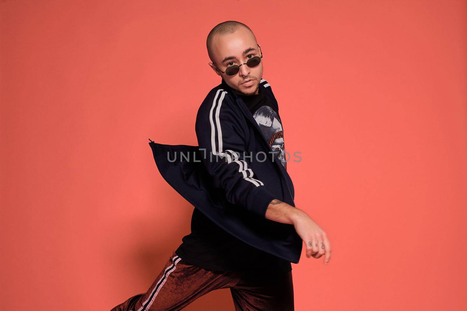 Close-up studio shot of a adult tattoed bald fellow in sunglasses, with a pirsing ring in his nose, wearing black trendy t-shirt with print and sport suit, fooling around while posing against a pink background with copy space. People, style and fashion concept. 90s style